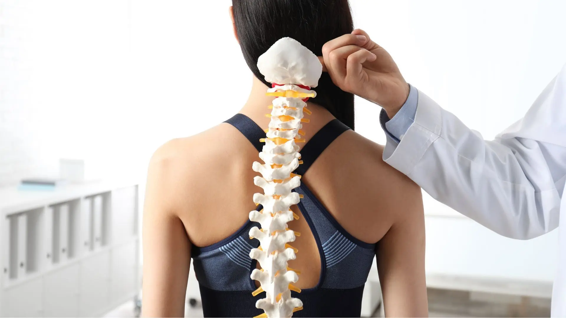 Doctor-Demonstrating-Spinal-Anatomy-in-Medical-Office A doctor in a white coat points at a spine model next to a woman's back, demonstrating spinal anatomy. The setting is a bright medical office.